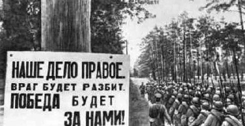 卫国战争（1941年6月22日—1945年5月9日）时期的黑白照片，一列苏联士兵行军经过写着“我们的事业是正义的，敌人必败，胜利属于我们”的标语牌